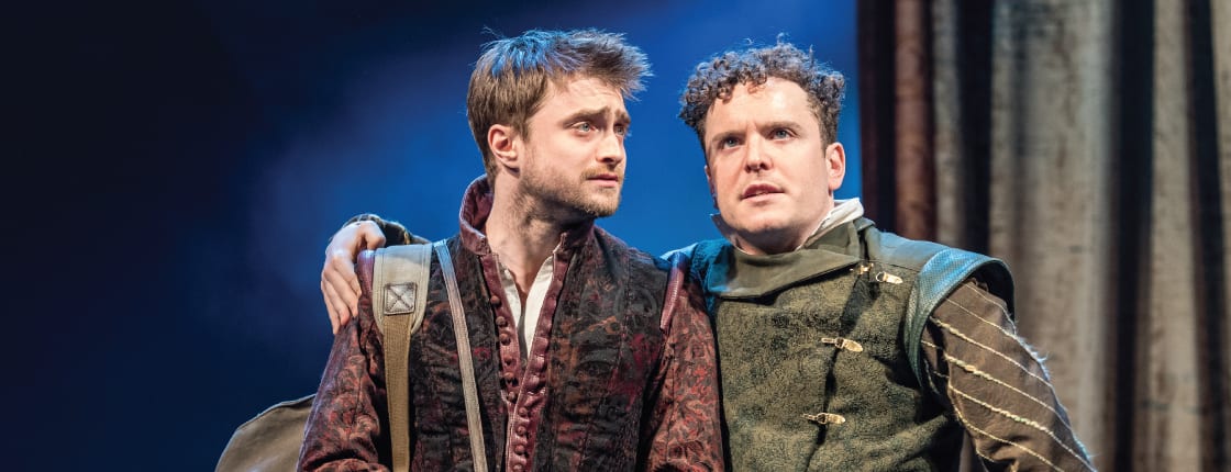 Rosencrantz and Guildenstern on stage with blue background, face forward with arms around each others shoulders